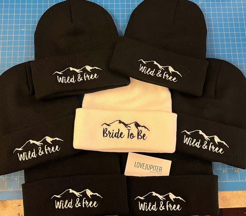 Puede incluir: Seis gorros de punto negros con bordado blanco de monta&ntilde;a y texto. Un gorro es blanco con bordado negro de monta&ntilde;a y texto. El texto en los gorros negros dice "Wild & Free". El texto en el gorro blanco dice "Bride To Be".