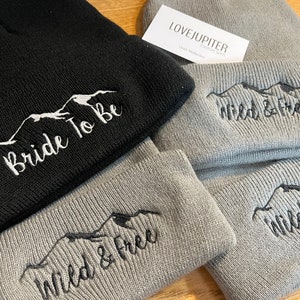 Puede incluir: Dos gorros de punto grises y uno negro con bordado de monta&ntilde;a y texto. Los gorros grises tienen "Wild & Free" bordado en ellos. El gorro negro tiene "Bride To Be" bordado en &eacute;l. Los gorros est&aacute;n doblados y apilados sobre una superficie de madera.