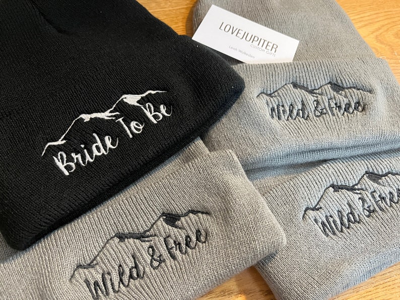 Puede incluir: Dos gorros de punto grises y uno negro con bordado de monta&ntilde;a y texto. Los gorros grises tienen "Wild & Free" bordado en ellos. El gorro negro tiene "Bride To Be" bordado en &eacute;l. Los gorros est&aacute;n doblados y apilados sobre una superficie de madera.