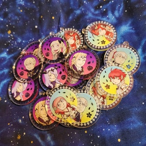 May include: A collection of round, colorful, clear acrylic charms featuring anime characters. Each charm has a gold border and a white background with a different character on each charm.