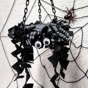 May include: A black and white striped spider-shaped hanging toy with black fuzzy legs and googly eyes. The toy is hanging from a black chain against a white background with a black spiderweb.