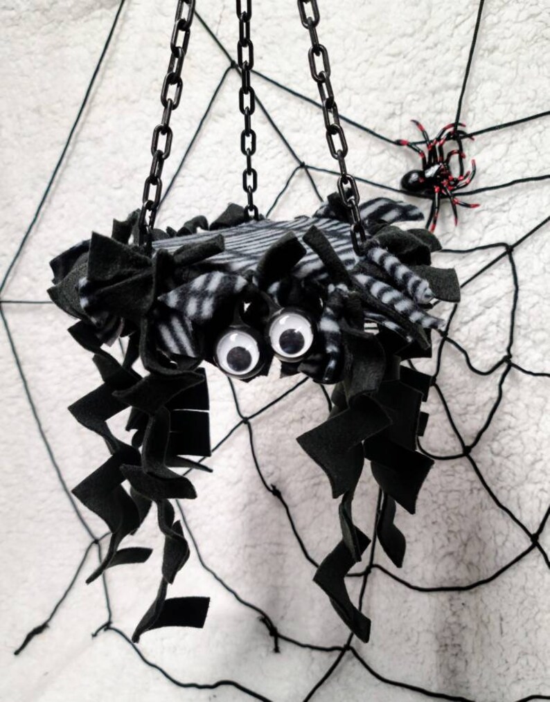 May include: A black and white striped spider-shaped hanging toy with black fuzzy legs and googly eyes. The toy is hanging from a black chain against a white background with a black spiderweb.
