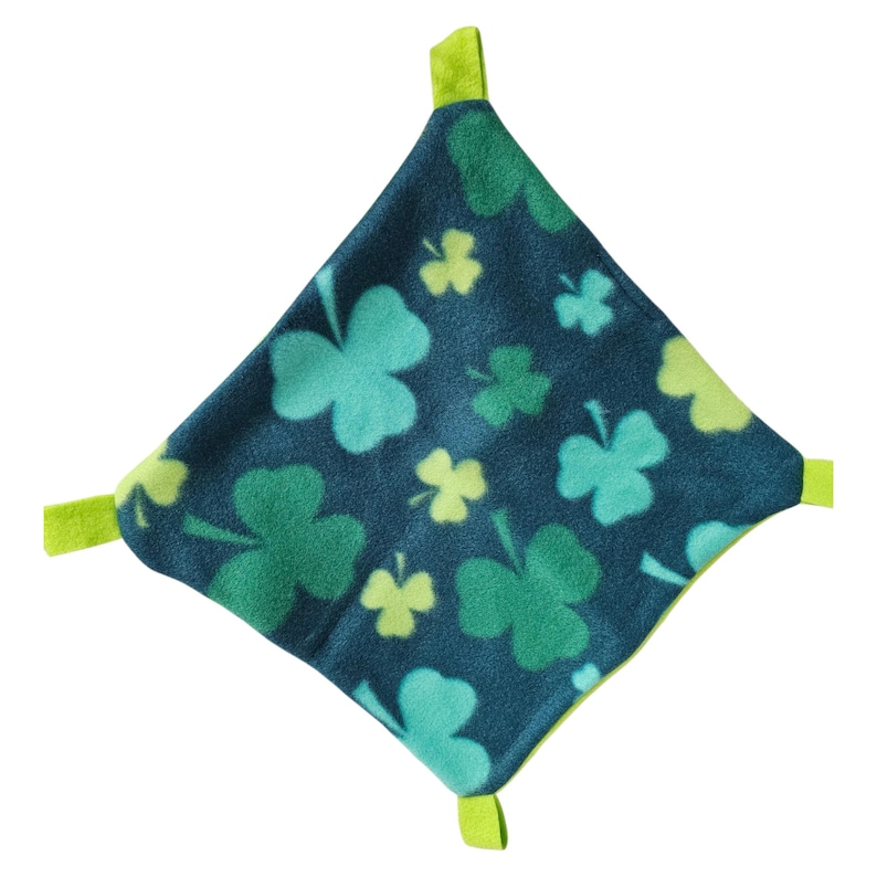 May include: A square, dark teal fleece blanket with a repeating pattern of green and light green shamrocks. The blanket has four lime green tabs at each corner.