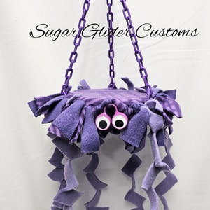 May include: Purple hanging toy for sugar gliders, featuring a plush platform with googly eyes and multiple dangling strands of fabric.