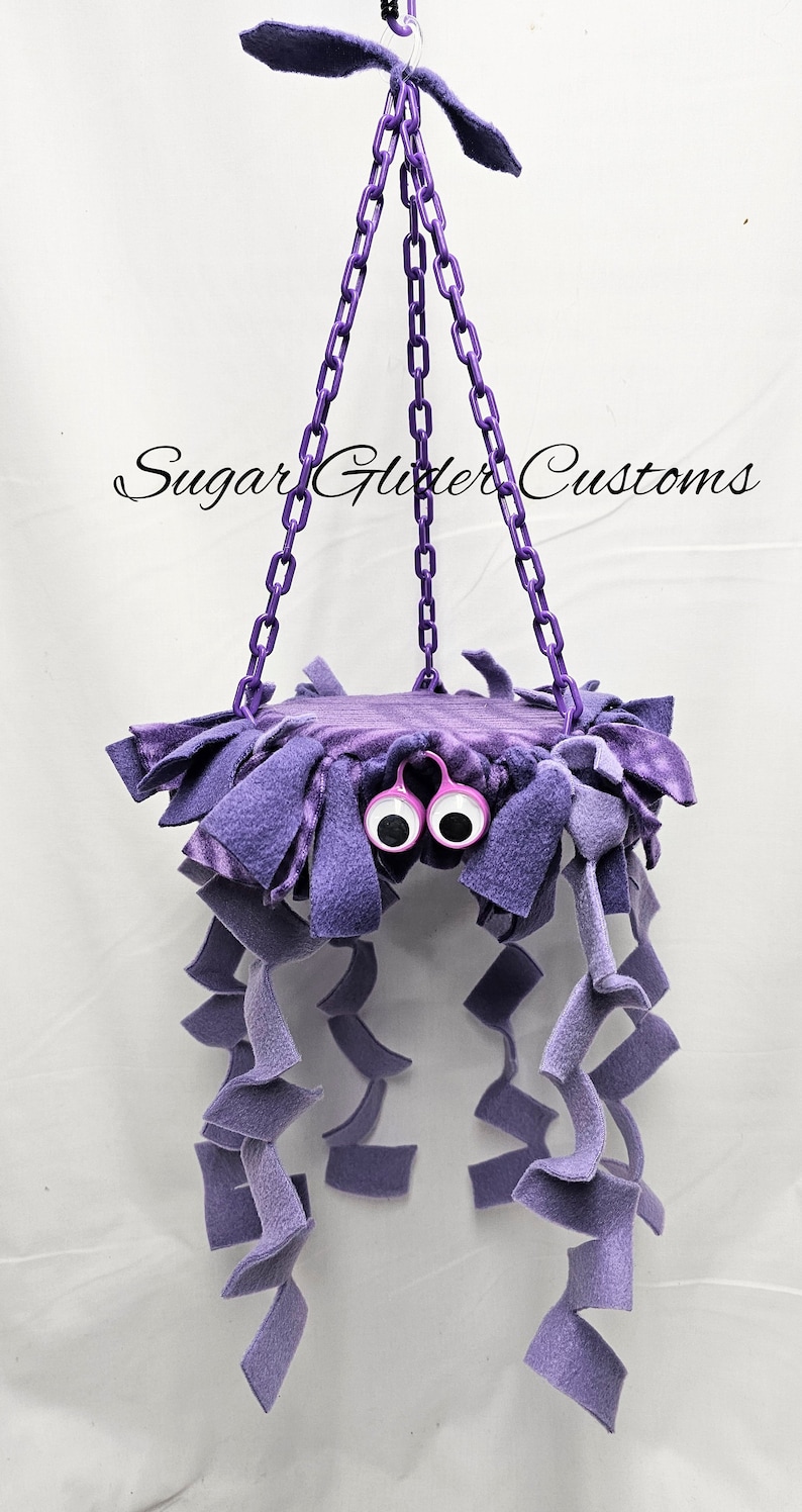 May include: Purple hanging toy for sugar gliders, featuring a plush platform with googly eyes and multiple dangling strands of fabric.