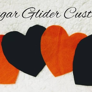 May include: Four felt heart shapes in orange and black are arranged on a white, textured surface. The text "Sugar Glider Customs" is written in cursive at the top of the image. The hearts are evenly spaced.