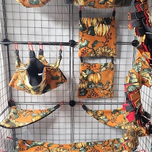 May include: A set of  sugar glider cage accessories in a fall pumpkin and sunflower print. The set includes a hammock, a hanging pouch, a hanging bed, a fleece toy, and a small pouch. The accessories are all made of soft fleece fabric and are designed to provide a comfortable and stimulating environment for sugar gliders. The set also includes a small bag of colorful plastic toys. The text "Sugar Glider Customs" is visible in the image.