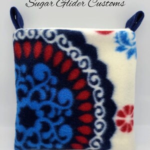 May include: A square fleece pouch with two dark blue fabric loops at the top corners. The fabric features a white background with a large, circular mandala-style pattern in shades of dark blue, bright blue, and red. Text reads Sugar Glider Customs.
