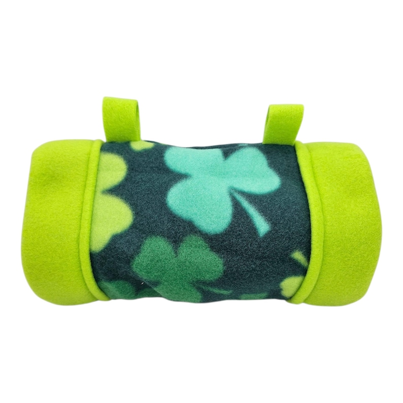 May include: A cylindrical pet bed made of green and dark green fleece. The center section features a pattern of shamrocks in shades of green. The ends and two small loops are a bright green color.