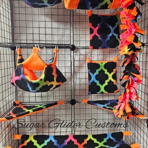 May include: A selection of sugar glider cage accessories displayed in a wire cage. The items include hammocks, tunnels, and hanging beds. The accessories have a black background with a colourful, geometric pattern and bright orange trim. The text "Sugar Glider Customs" is visible.