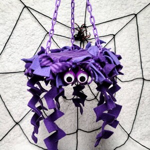 May include: Purple spider-shaped hanging toy with googly eyes and long, curly purple fabric tentacles. The toy is hanging from a chain against a white background with a black spiderweb.