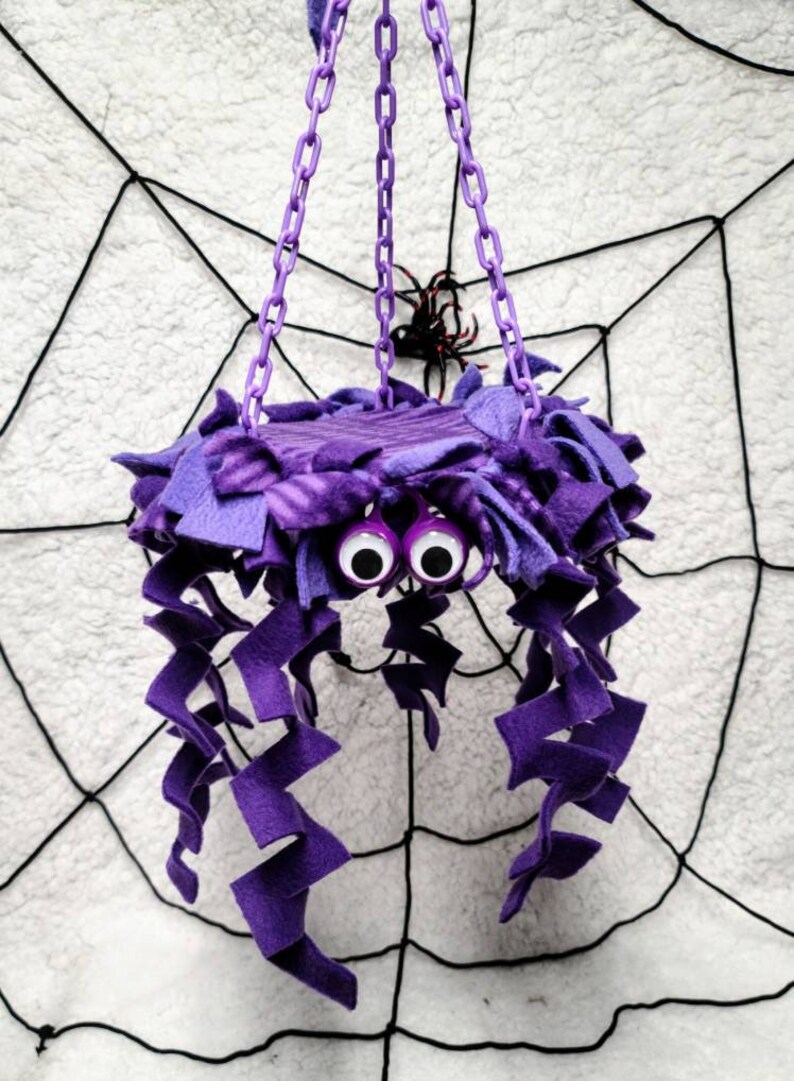 May include: Purple spider-shaped hanging toy with googly eyes and long, curly purple fabric tentacles. The toy is hanging from a chain against a white background with a black spiderweb.