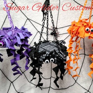 May include: Three handmade spider-shaped hanging toys for small pets. The toys are made of felt and have googly eyes. The spiders are purple, black and white, and orange.