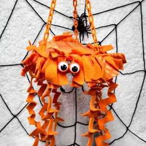 May include: Orange and white spider-shaped hanging toy with googly eyes and long orange fabric tentacles. The toy is hanging from a chain against a white background with a black spiderweb.