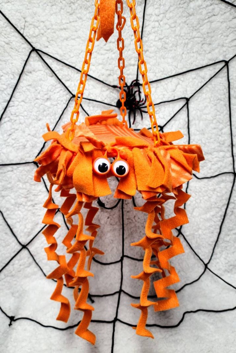 May include: Orange and white spider-shaped hanging toy with googly eyes and long orange fabric tentacles. The toy is hanging from a chain against a white background with a black spiderweb.