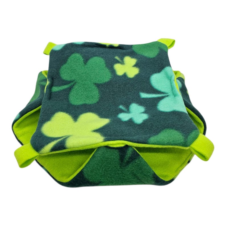 May include: A green and lime green fleece pet bed with a shamrock pattern. The bed is square-shaped with a contrasting lime green interior and trim. The shamrocks are in varying shades of green.