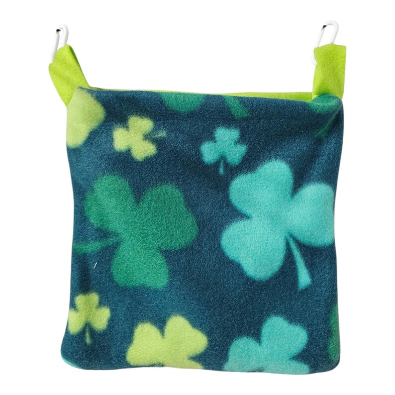 May include: A small, dark blue fleece pet bed with a repeating pattern of green and light green shamrocks. The bed has lime green trim and two metal clips for hanging.