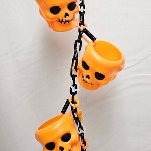May include: A Halloween-themed toy for birds, featuring three orange skull-shaped cups hanging from a black chain with a white spider and a glow-in-the-dark skeleton hand.