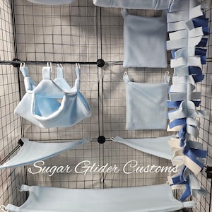 May include: A selection of light blue fleece pet accessories, including hammocks, hanging beds, and a fringed toy, displayed inside a wire cage. The accessories are designed for small pets and are made of soft, comfortable material.