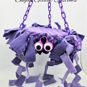 May include: Purple hanging toy for sugar gliders, with a soft, plush fabric and dangling strips. The toy has large googly eyes and a purple chain.