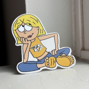 Può includere: Un adesivo a fumetti di una ragazza con i capelli biondi, che indossa un top giallo con il logo dei Nashville Predators, pantaloni blu e scarpe gialle. La ragazza è appoggiata sulla mano, con un'espressione pensierosa.