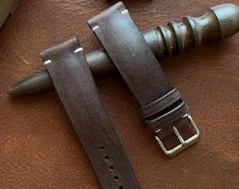 May include: A pair of brown leather watch straps with white stitching and a gold-colored buckle. A Seiko watch with a green face is in the background. The watch straps are on a brown surface.