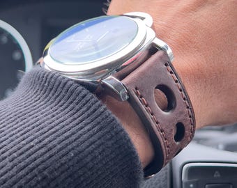 Könnte beinhalten: Eine silberne Armbanduhr mit einem braunen Lederarmband mit großen kreisförmigen Aussparungen. Die Uhr hat ein dunkles Zifferblatt und wird am Handgelenk getragen, mit einem schwarzen Pulloverärmel sichtbar.