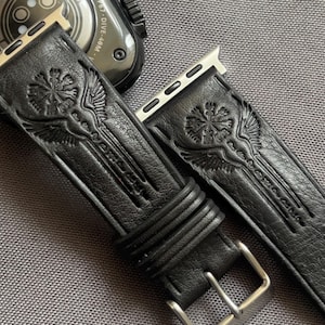 May include: Black leather watch straps with an embossed design featuring a central emblem with wings. The straps have a silver buckle and are designed for a smartwatch. The straps are shown on a grey surface.