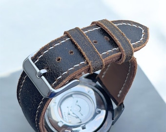 May include: Close-up of a wristwatch with a distressed brown leather strap. The strap is stitched with white thread and has a silver buckle. The watch face is visible, with a silver case and a clear back.