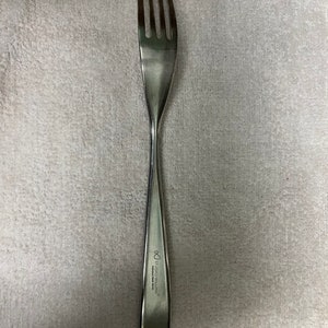Puede incluir: Un tenedor de acero inoxidable con cuatro púas. El mango es liso y se estrecha hacia el extremo, con el texto "STUDIO WILLIAM" y un logotipo grabado. El tenedor está diseñado para comer.