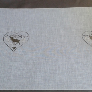 Puede incluir: Camino de mesa blanco con dos corazones bordados que representan un ciervo y montañas. Los corazones están bordeados de negro y tienen un pequeño corazón en el centro.