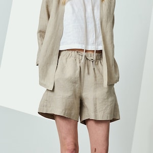 Può includere: Una donna indossa un completo in lino beige, composto da un cappello a secchiello, un blazer, pantaloncini e un top bianco. Indossa scarpe da ginnastica bianche. Lo sfondo è un muro bianco con forme geometriche.