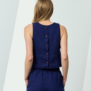 Pode incluir: Um conjunto de duas peças de linho azul marinho. O top sem mangas tem uma fileira de botões nas costas. As calças de perna larga têm bolsos. O conjunto é feito de um tecido texturizado.