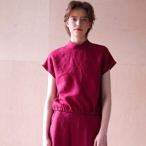 May include: A magenta-colored dress with a high neckline and short sleeves. The dress has a cinched waist and a long, flowing skirt. The person is wearing brown heeled sandals. The background is a light wood panel.