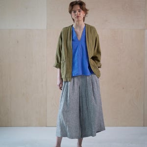 May include: A woman wearing a casual outfit. The ensemble includes a blue V-neck top, a green jacket, and gray wide-leg pants. The outfit is completed with black flats. The background is a light wood panel.