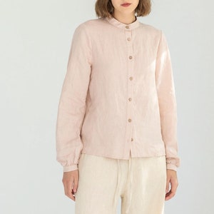 May include: A light pink linen shirt with a mandarin collar and wooden buttons is paired with cream-colored linen pants. The pants have a relaxed fit and a drawstring waist. The outfit is completed with black sandals.