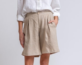 May include: A woman in a white linen shirt and beige linen shorts. The shorts are high-waisted and pleated. She is wearing brown sandals with black soles. The outfit is casual.