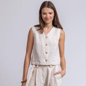 May include: A beige linen two-piece outfit. The sleeveless top has a V-neck and wooden buttons. The high-waisted, wide-leg pants have a matching fabric belt. The outfit is made of a lightweight, textured fabric, perfect for warm weather.