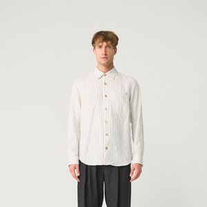 Op de afbeelding: Een wit overhemd met grijze verticale strepen en houten knopen, gecombineerd met een donkergrijze plooibroek en zwarte schoenen. De outfit wordt gepresenteerd tegen een witte achtergrond, wat een minimalistische esthetiek benadrukt.