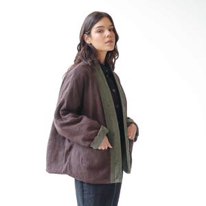May include: A brown jacket with green lining and trim. The jacket is worn over a black shirt and paired with black trousers and black boots. The jacket has a relaxed fit and a casual style.