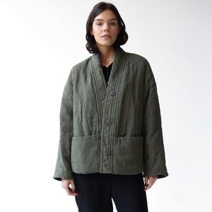 May include: An olive green quilted jacket with a relaxed fit, featuring a button-up front and two front pockets. The model is wearing black pants and black boots. The jacket appears to be made of a soft, textured fabric.