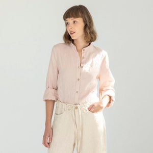 May include: A light pink linen shirt with a mandarin collar and button-down front is paired with cream-colored wide-leg linen pants. The pants have a drawstring waist. The outfit is completed with beige sneakers.