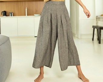 May include: A woman wearing a mustard yellow sleeveless top and gray wide-leg linen pants. The pants have a high waist and pleats. The outfit is casual and comfortable, suitable for everyday wear. The background is a modern kitchen with white cabinets and wooden accents.