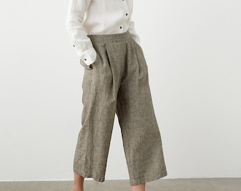 May include: A woman wearing a white button-down shirt with black buttons and cuffs, paired with wide-leg, cropped, gray-brown pants. She is also wearing brown leather clogs. The outfit is set against a plain white background.