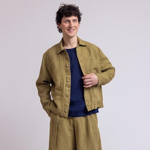 May include: An olive green button-down jacket with a collar and two front pockets, paired with matching olive green knee-length shorts featuring a tie waist, worn over a solid navy blue short-sleeved shirt and light beige lace-up sneakers.