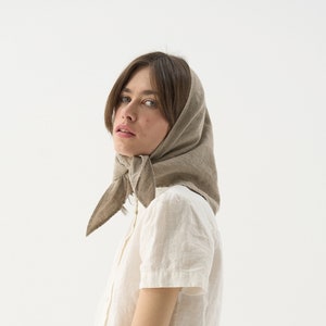 Può includere: Una donna indossa un foulard di lino beige chiaro, una camicia bianca abbottonata e pantaloni di lino verde oliva. La donna tiene in mano un cesto di fiori secchi e verde. L'outfit è semplice ed elegante.