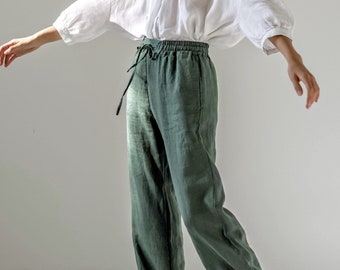 May include: A woman wearing a white linen blouse with wide sleeves and olive green linen pants. The pants have a drawstring waist. Black sandals complete the outfit. The clothing appears to be lightweight and suitable for warm weather.
