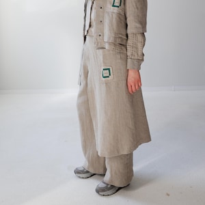 Puede incluir: Un conjunto de lino beige claro que consta de una camisa de manga larga, pantalones de pernera ancha y una falda larga. La camisa y la falda tienen parches cuadrados verdes. Se lleva una bufanda a juego. El conjunto se completa con zapatillas grises.