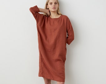 May include: A knee-length, rust-colored dress with a round neckline and three-quarter length sleeves. The garment features a loose, straight-cut silhouette and is paired with simple black slide sandals on a neutral floor surface.