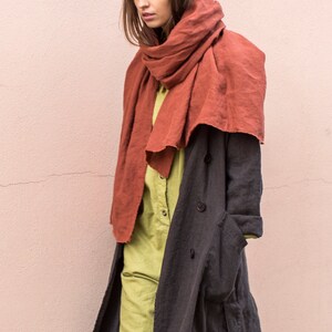 May include: A woman wearing a rust-colored scarf, a light green jumpsuit, and a dark gray long coat. The coat has large buttons and pockets. The outfit is a stylish ensemble.